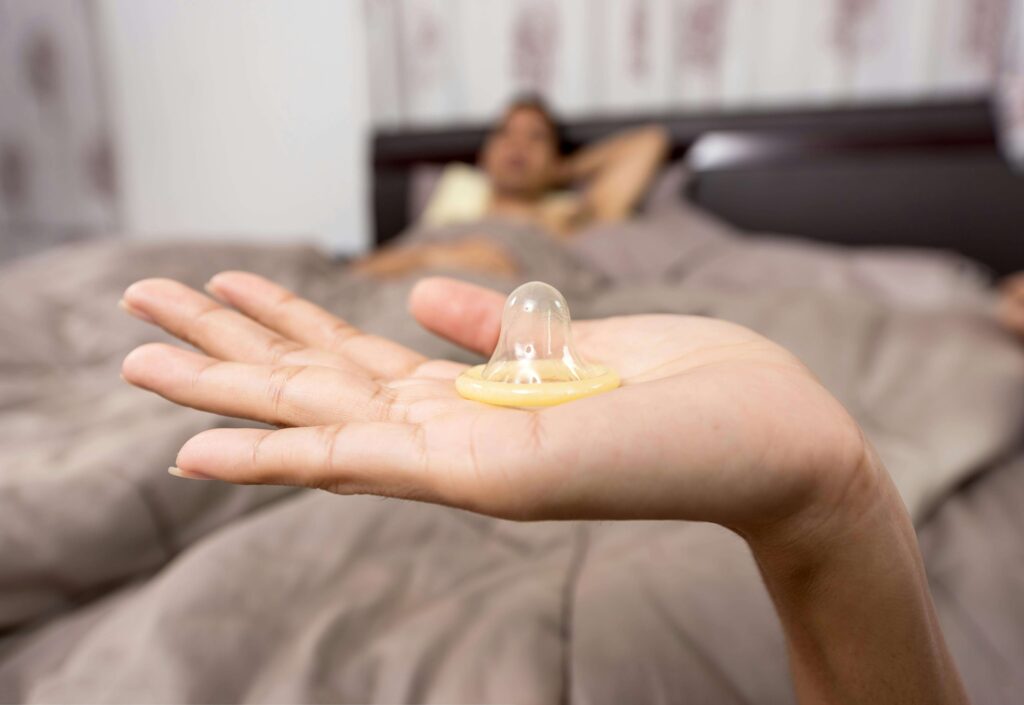 an escort holding a condom in front of a bed with a client